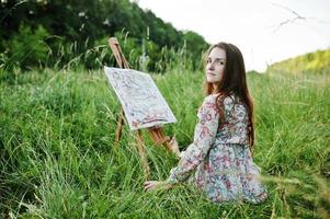 retrato de uma linda jovem feliz em lindo vestido sentado na grama e pintando em papel com aquarelas. foto