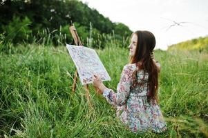 retrato de uma linda jovem feliz em lindo vestido sentado na grama e pintando em papel com aquarelas. foto