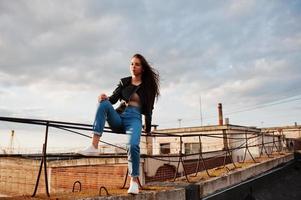 retrato de uma linda jovem de jaqueta de couro preta, jeans e tênis sentado em corrimãos no telhado com vista pitoresca de um parque. foto