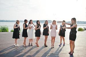 grupo de 8 meninas usam preto e 2 noivas na festa de despedida contra praia ensolarada bebendo champanhe. foto