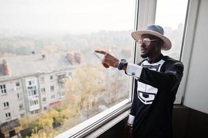 homem africano rico olhando para janela panorâmica em sua cobertura. retrato de homem negro bem sucedido no chapéu interior. foto