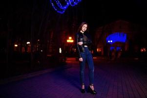 retrato noturno do modelo de menina usar óculos, jeans e jaqueta de couro contra guirlanda de luzes azuis da rua da cidade. foto