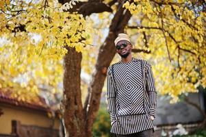 retrato de homem afro-americano elegante preto no chapéu e óculos de sol contra o fundo de outono ensolarado outono. pessoas ricas da áfrica em trajes tradicionais. foto