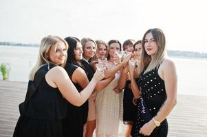 grupo de 8 meninas usam preto e 2 noivas na festa de despedida contra praia ensolarada bebendo champanhe. foto