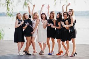 grupo de 8 meninas usam preto e 2 noivas na festa de despedida contra praia ensolarada bebendo champanhe. foto