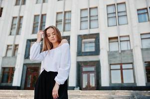 retrato de uma jovem fabulosa bem sucedida na blusa branca e calças pretas largas posando nas escadas com um enorme edifício branco no fundo. foto