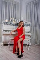 mulher linda elegante no vestido de noite vermelho longo sentado no estúdio contra piano com velas decoração de ano novo. foto