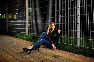 retrato noturno do desgaste do modelo de menina em jeans e jaqueta de couro contra cerca de ferro. foto