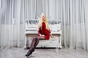 menina loira elegante usar vestido vermelho posou contra piano no estúdio. foto