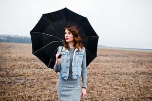 retrato de menina morena encaracolada na jaqueta jeans com guarda-chuva preto no campo. foto