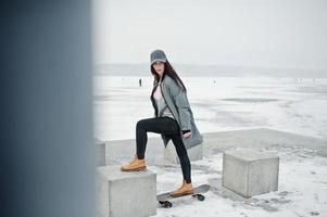 menina morena elegante boné cinza, estilo casual de rua com skate em dia de inverno. foto