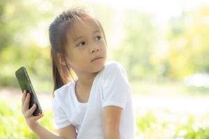 linda criança asiática sorrindo usando telefone celular inteligente no jardim, criança tem paixão jogando jogo digital em smartphone na rede de internet no parque com conceito feliz, estilo de vida e tecnologia. foto
