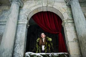 menina morena com casaco de pele verde contra velho arco com colunas e cortinas vermelhas. foto