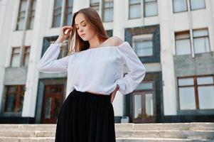 retrato de uma jovem fabulosa bem sucedida na blusa branca e calças pretas largas posando nas escadas com um enorme edifício branco no fundo. foto