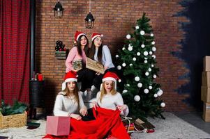 quatro amigas fofas usam blusas quentes, calças pretas e chapéus de papai noel contra a árvore de ano novo com decoração de natal no estúdio. foto