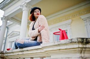garota ruiva de óculos e chapéu com bolsa vermelha posou perto de casa vintage. foto