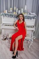 mulher linda elegante no vestido de noite vermelho longo sentado no estúdio contra piano com velas decoração de ano novo. foto