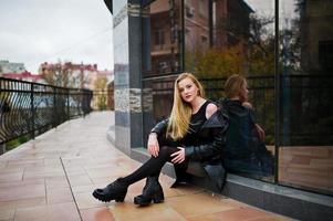 menina elegante loira com casaco de couro preto longo posou contra a grande janela do edifício. foto