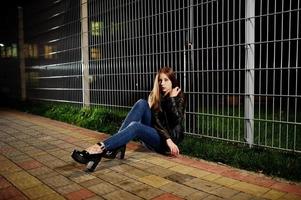 retrato noturno do desgaste do modelo de menina em jeans e jaqueta de couro contra cerca de ferro. foto