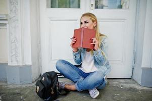 menina loira usar jeans com mochila posou contra a porta velha com diário e escrever algo. foto