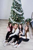 quatro amigas fofas usam blusas quentes, calças pretas contra a árvore de ano novo com decoração de natal no quarto branco. foto