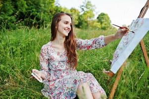 retrato de uma linda jovem feliz em lindo vestido sentado na grama e pintando em papel com aquarelas. foto