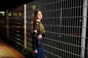 retrato noturno do desgaste do modelo de menina em jeans e jaqueta de couro contra cerca de ferro. foto