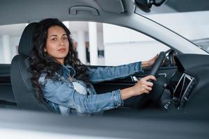 tempo para um passeio. linda garota com cabelo preto tentando seu novo carro caro no salão de automóveis foto