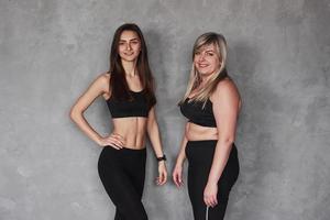 duas amigas com diferentes tipos de corpo e em roupas esportivas em pé no estúdio com fundo cinza foto