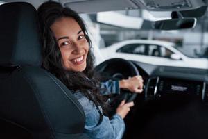 sorriso dentuço. linda garota com cabelo preto tentando seu novo carro caro no salão de automóveis foto