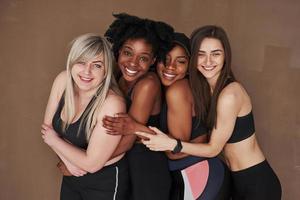 inclinando-se para a frente deles. grupo de mulheres multiétnicas em pé no estúdio contra fundo marrom foto