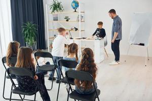 olhando para o menino com microscópio que está em cima da mesa. grupo de alunos de crianças em sala de aula na escola com professor foto