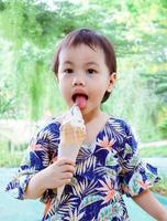 Menina asiática de 3 anos de idade, bonitinha, enfiando a língua para fora e lambendo sorvete ao ar livre. foto