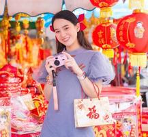 mulher bonita asiática em um qipao azul-cinza está segurando um saco de pano com a palavra feliz em chinês e segurando uma câmera rosa. fundos são objetos auspiciosos para decorar casas chinesas. foto