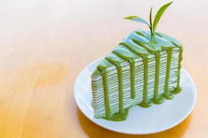 bolo de crepe de chá verde matcha que por cima com molho matcha e decorado com folhas de chá verde fresco colocado em um prato branco sobre uma mesa de madeira.
