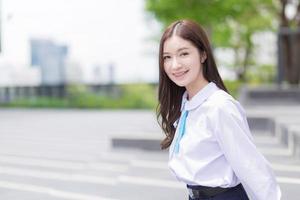 linda estudante do ensino médio asiático no uniforme escolar com aparelho nos dentes fica e sorri com confiança enquanto ela olha para a câmera alegremente com a cidade ao fundo. foto