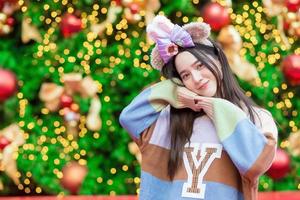 uma linda mulher asiática em um suéter colorido fica sorrindo e feliz na frente da árvore de natal com bokeh como pano de fundo no tema para comemorar o natal e feliz ano novo foto