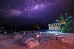 noite de cinema na praia estrelada da ilha tropical. vista panorâmica incrivelmente calma e descontraída do cinema ao ar livre com a via láctea e palmeiras com luz suave de velas. férias em família de verão, estilo de vida de luxo foto