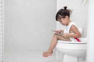 criança sentada no vaso sanitário segurando o tablet. conceito de smartphone viciado em criança foto