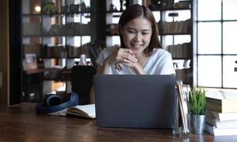 estudante asiática de graduação adolescente sorridente estuda na biblioteca com livros de laptop fazendo pesquisas on-line para cursos, fazendo anotações para trabalhos de casa de ensaio, conceito de e-learning de educação on-line foto