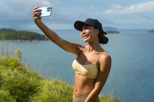 linda mulher asiática em roupas esportivas tirando foto no pico da montanha à beira-mar após o conceito de trekking, viagens e ecoturismo.