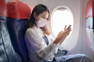 jovem mulher usando máscara facial está usando smartphone a bordo, nova viagem normal após o conceito de pandemia covid-19 foto