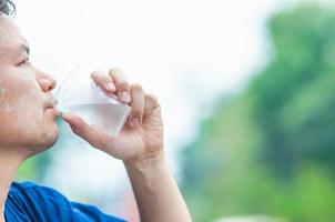 homem do norte da Tailândia bebe água fresca e fria em vidro plástico durante a atividade ao ar livre de participação em dia muito quente, vestindo camisa de estilo do norte da Tailândia foto