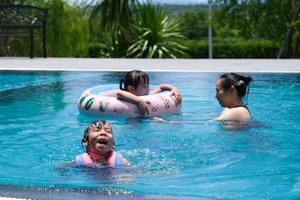 irmãs bonitas estão nadando em coletes salva-vidas com a mãe na piscina em um dia ensolarado. família feliz, mãe e filhas brincando na piscina. conceito de estilo de vida de verão.