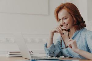 modelo feminino satisfeito posa no espaço de coworking, segura óculos, sorri positivamente, usa conexão de internet sem fio, lê informações, tem comunicação online, trabalha na criação de projeto web foto