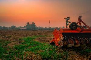 máquina de repressão cumeeira. fornecimento para trator. máquinas agrícolas na fazenda agrícola. fazenda de arroz de manhã com céu vermelho do nascer do sol. natureza das terras agrícolas. cabana de agricultor e poste elétrico em terras agrícolas. foto