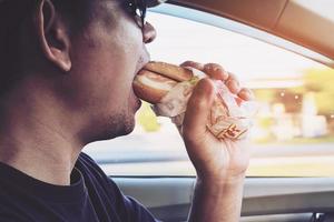 homem está comendo perigosamente cachorro-quente e bebida gelada enquanto dirige um carro foto