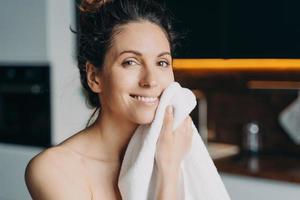 menina europeia está limpando o rosto com a toalha após a lavagem. jovem morena toma banho em casa. foto