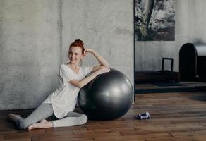 linda mulher ruiva esportiva sentada no chão de madeira com grande fitball prata foto