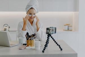 blogueira muito feliz grava streaming de vídeo para seu blog dá tutorial sobre como aplicar adesivos de beleza dá conselhos sobre cuidados com a pele vestida de roupão de banho olha para poses de câmera na mesa na cozinha foto